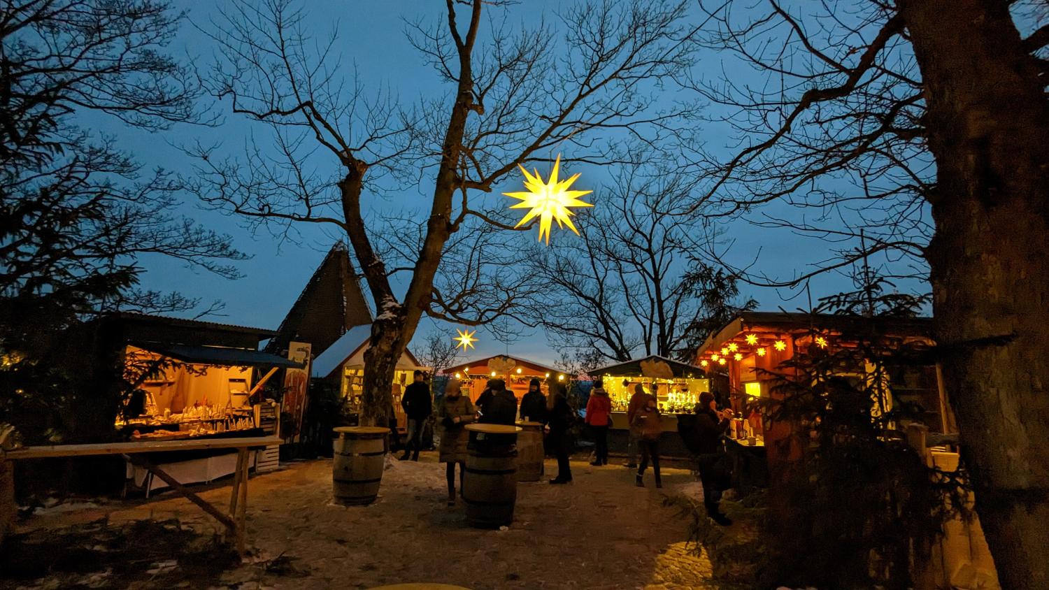 Weihnachtsmarkt Das Foto zeigt einen stimmungsvollen Weihnachtsmarkt in der Abenddämmerung mit beleuchteten Holzständen, kahlen Bäumen und großen leuchtenden Sternen, die über den Besucherinnen und Besuchern hängen.