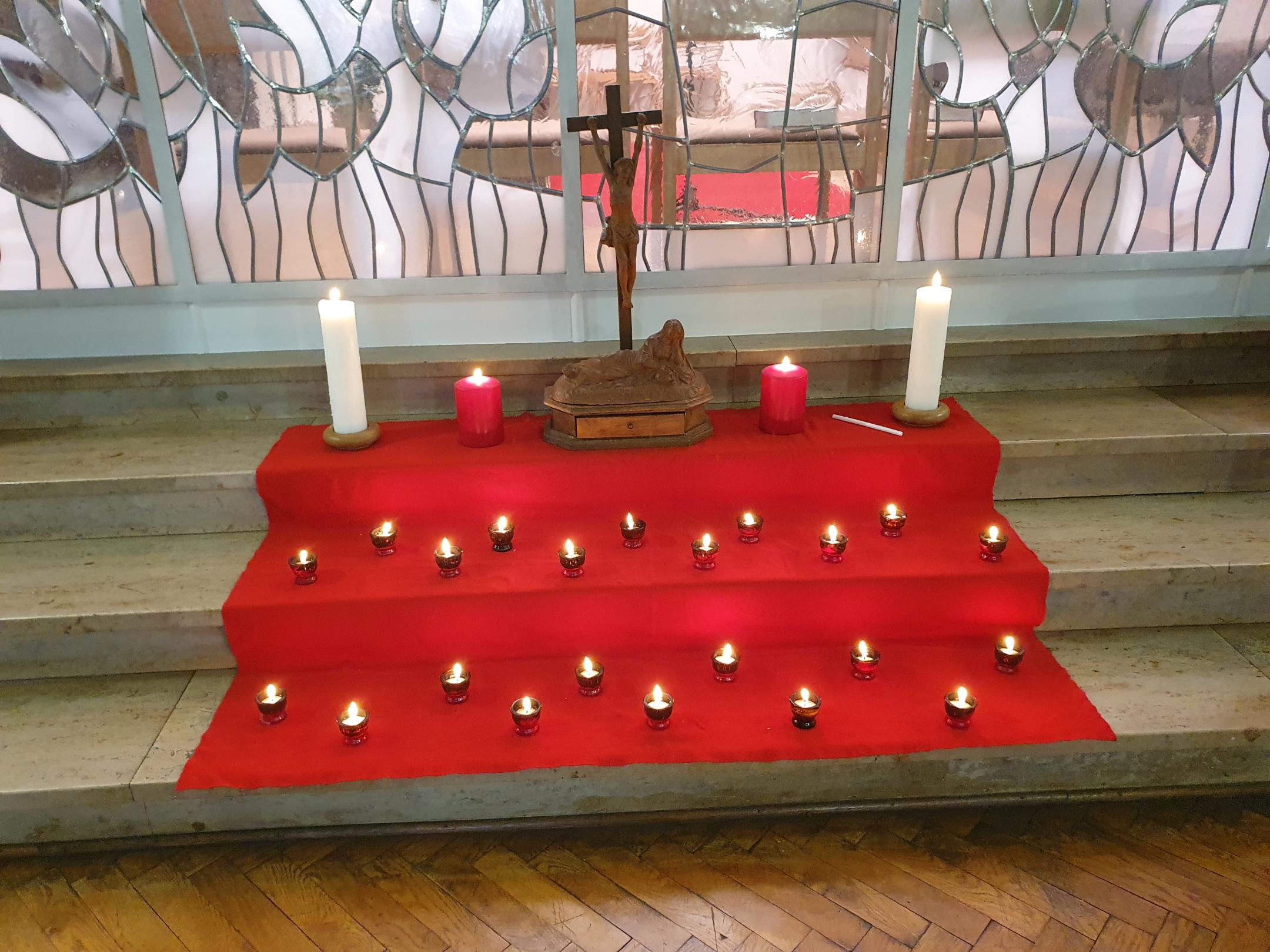 eine rote Decke auf drei Breiten Stufen, darauf steht oben ein Kreuz eingerahmt von großen Kerzen. Davor auf den Stufen darunter kleine Kerzen. Im Hintergrund ein Kirchenfenster