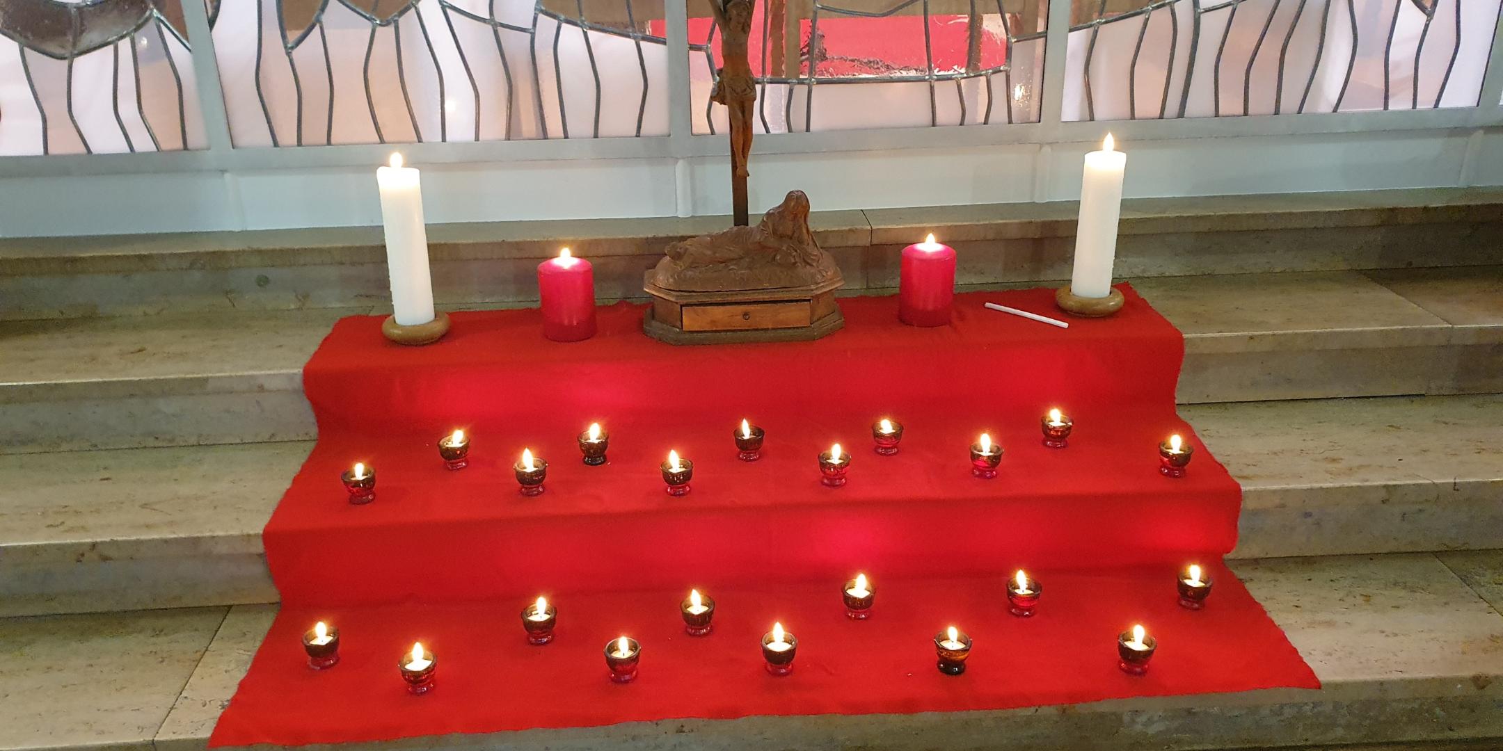 eine rote Decke auf drei Breiten Stufen, darauf steht oben ein Kreuz eingerahmt von großen Kerzen. Davor auf den Stufen darunter kleine Kerzen. Im Hintergrund ein Kirchenfenster
