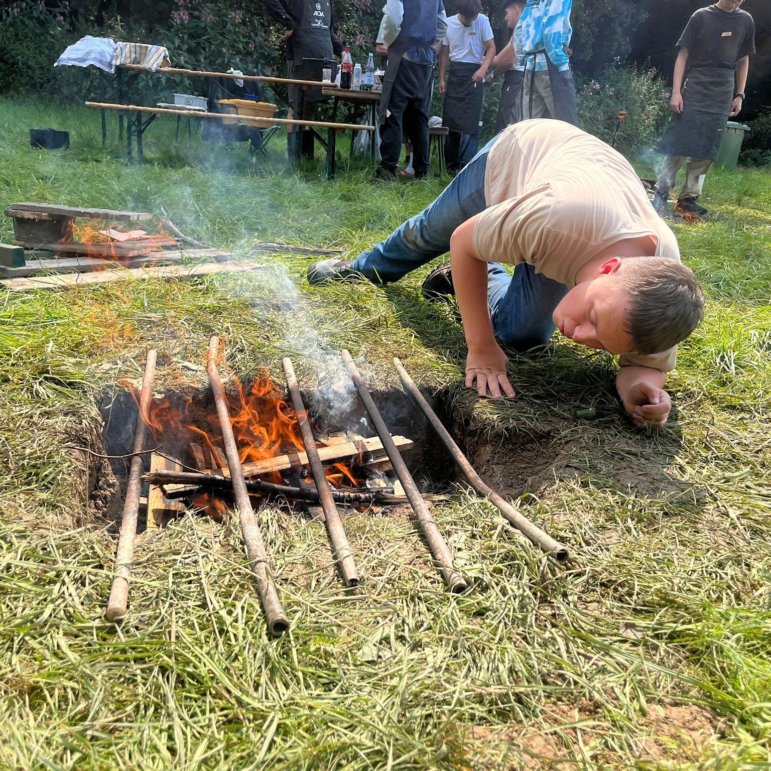 Kochgrube mit offenem Feuer in einer Wiese, ein Jugendlicher entfacht oder prüft es