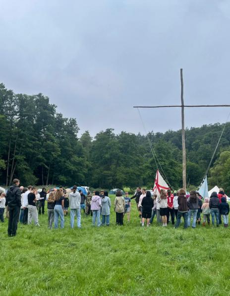 Im Fokus ist ein großes einfaches Kreuz aus zwei Baumstämmen in einem Zeltlager auf einer Wiese