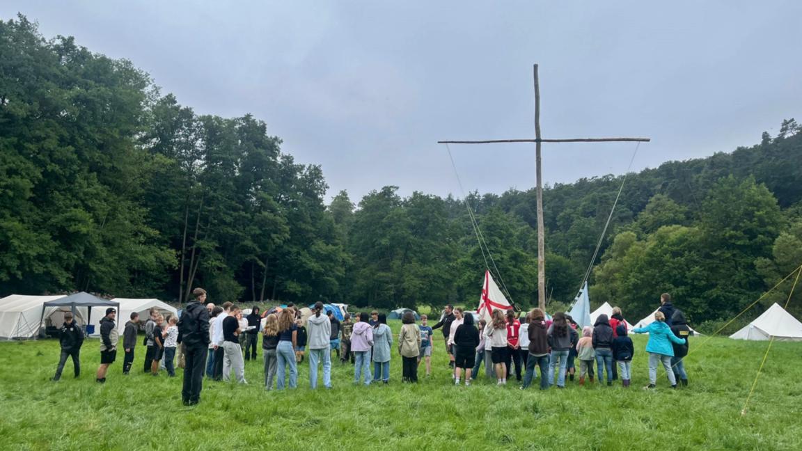Im Fokus ist ein großes einfaches Kreuz aus zwei Baumstämmen in einem Zeltlager auf einer Wiese