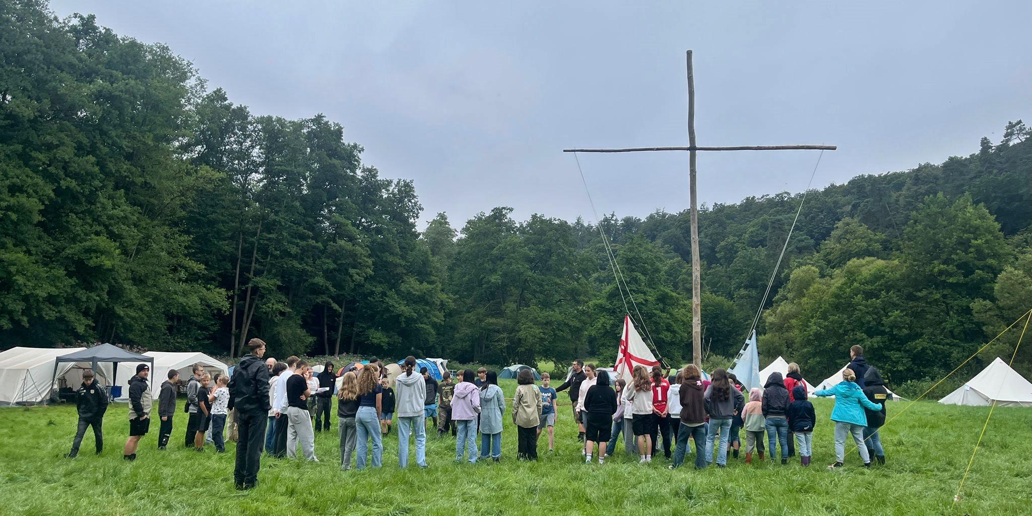 Im Fokus ist ein großes einfaches Kreuz aus zwei Baumstämmen in einem Zeltlager auf einer Wiese