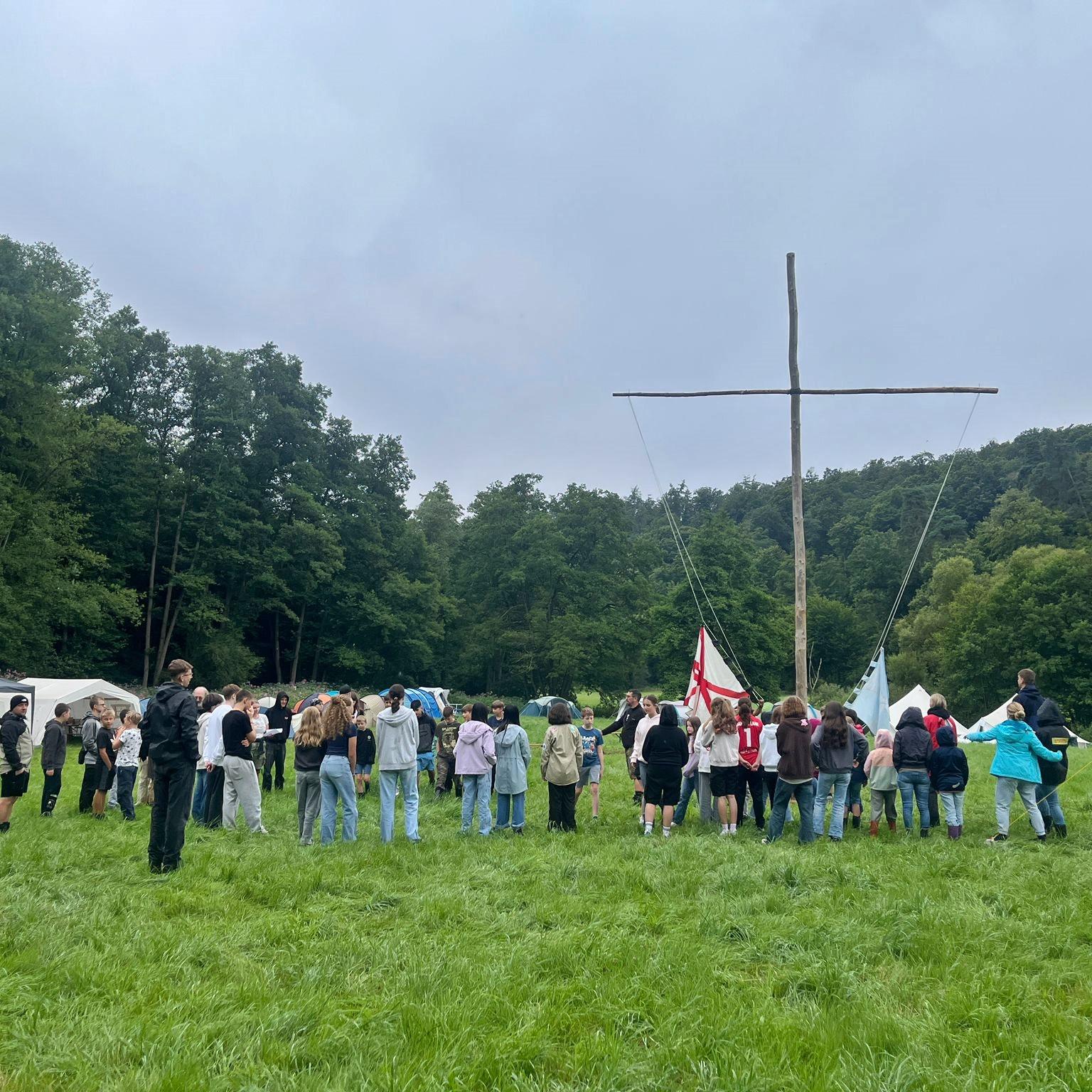 Im Fokus ist ein großes einfaches Kreuz aus zwei Baumstämmen in einem Zeltlager auf einer Wiese