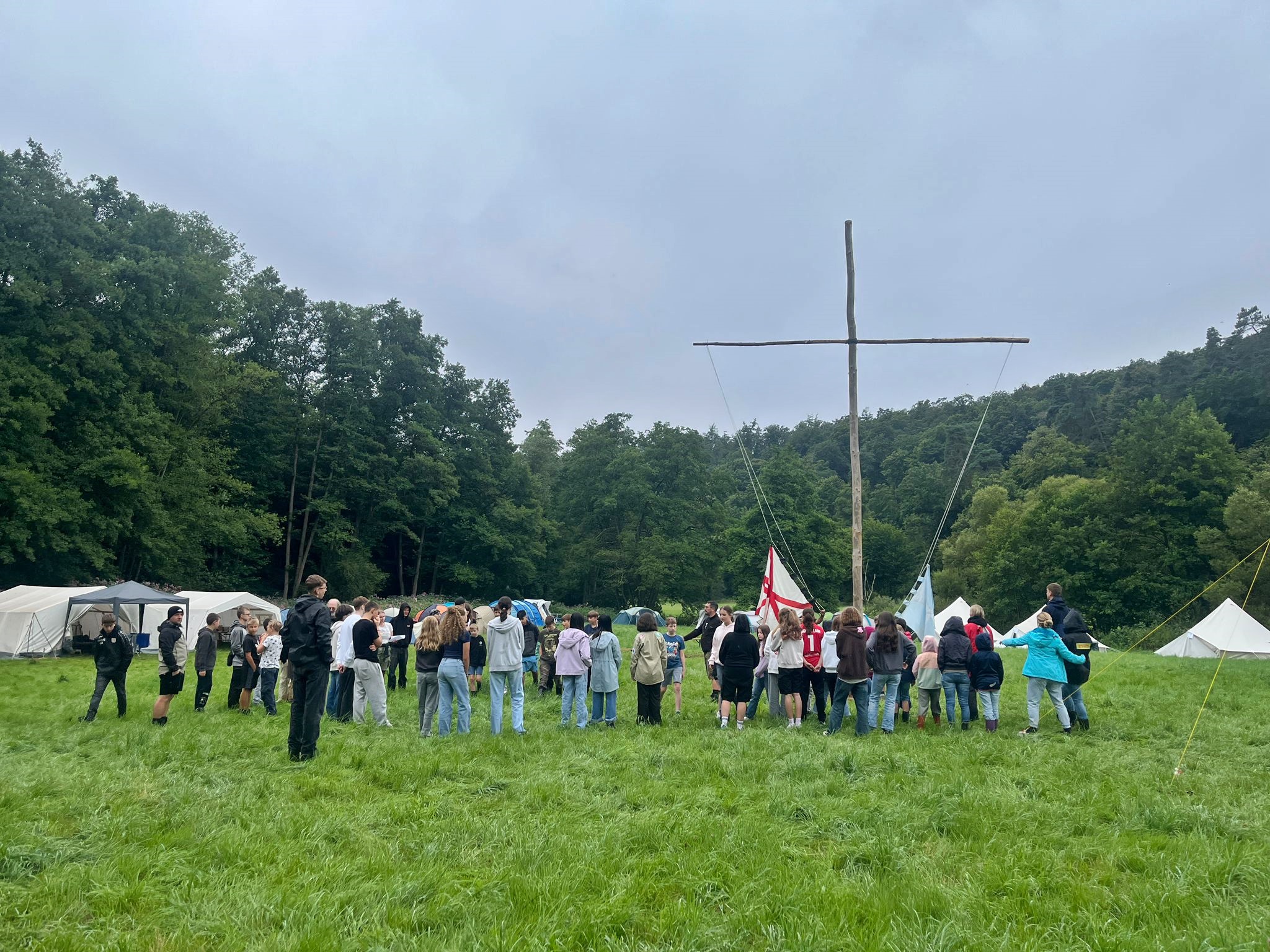 Im Fokus ist ein großes einfaches Kreuz aus zwei Baumstämmen in einem Zeltlager auf einer Wiese