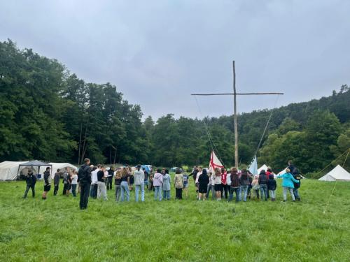 Im Fokus ist ein großes einfaches Kreuz aus zwei Baumstämmen in einem Zeltlager auf einer Wiese