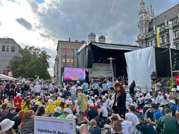 Blick auf eine Menschenmenge aus Kindern und Jugendlichen auf einem Platz in München, im Hintergrund eine Bühne mit einem Kreuz - Kinder- und Jugendchortag in München