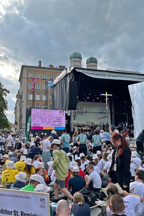 Blick auf eine Menschenmenge aus Kindern und Jugendlichen auf einem Platz in München, im Hintergrund eine Bühne mit einem Kreuz - Kinder- und Jugendchortag in München