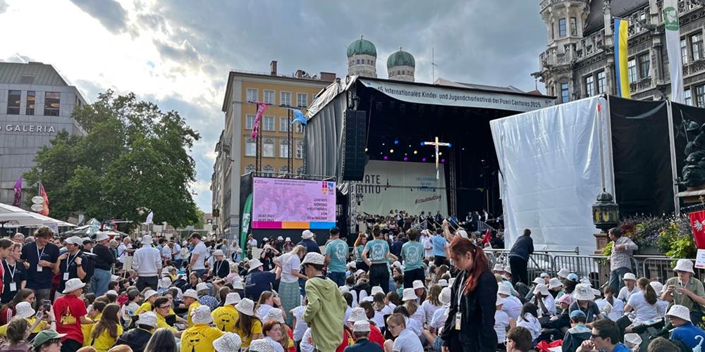 Blick auf eine Menschenmenge aus Kindern und Jugendlichen auf einem Platz in München, im Hintergrund eine Bühne mit einem Kreuz - Kinder- und Jugendchortag in München (c) Isabel Weiher Blick auf eine Menschenmenge aus Kindern und Jugendlichen auf einem Platz in München, im Hintergrund eine Bühne mit einem Kreuz - Kinder- und Jugendchortag in München