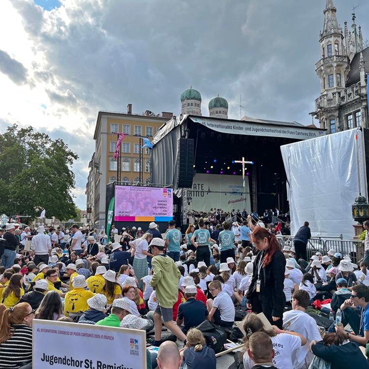 Blick auf eine Menschenmenge aus Kindern und Jugendlichen auf einem Platz in München, im Hintergrund eine Bühne mit einem Kreuz - Kinder- und Jugendchortag in München