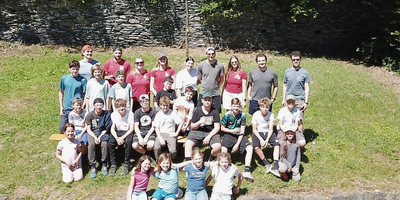 Eine Gruppe Jugendliche und Kinder sitzt auf einem grasbewachsenen Hügel und Natursteinmauer, dahinter ein selbstangefertigtes Kreuz (c) Leonie Castor für Pfarrei Koblenz St. Aposteln Eine Gruppe Jugendliche und Kinder sitzt auf einem grasbewachsenen Hügel und Natursteinmauer, dahinter ein selbstangefertigtes Kreuz
