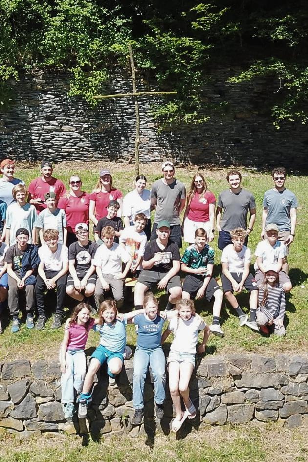 Eine Gruppe Jugendliche und Kinder sitzt auf einem grasbewachsenen Hügel und Natursteinmauer, dahinter ein selbstangefertigtes Kreuz