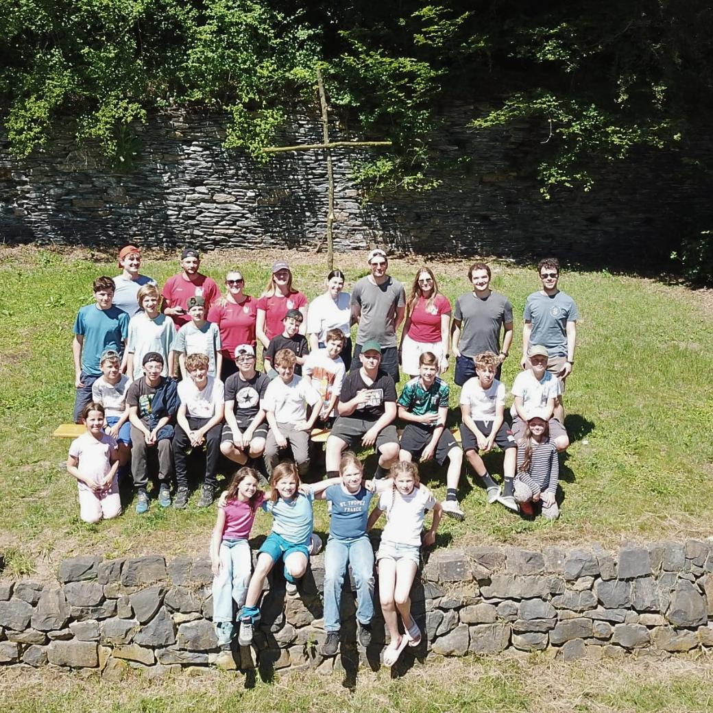 Eine Gruppe Jugendliche und Kinder sitzt auf einem grasbewachsenen Hügel und Natursteinmauer, dahinter ein selbstangefertigtes Kreuz