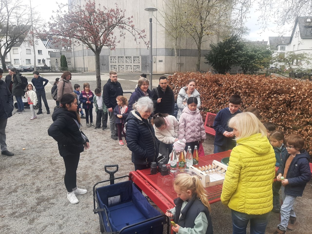 Familien, die auf einen Tisch zu gehen, auf dem Getränke und Schokohasen angeboten werden. Im Hintergrund ist die St. Franziskuskirche zu sehen