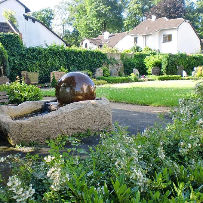 Blick auf einen Brunnen mit Sitzbank, im Hintergrund sind Grabstätten vor der Friedhofsmauer zu sehen