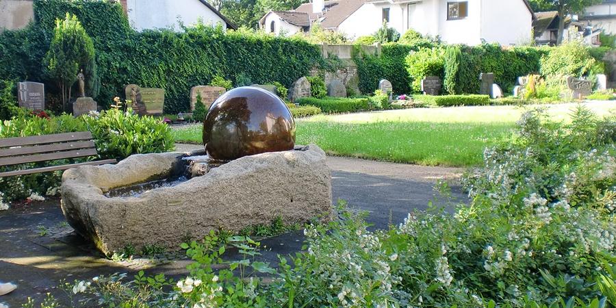 Blick auf einen Brunnen mit Sitzbank, im Hintergrund sind Grabstätten vor der Friedhofsmauer zu sehen