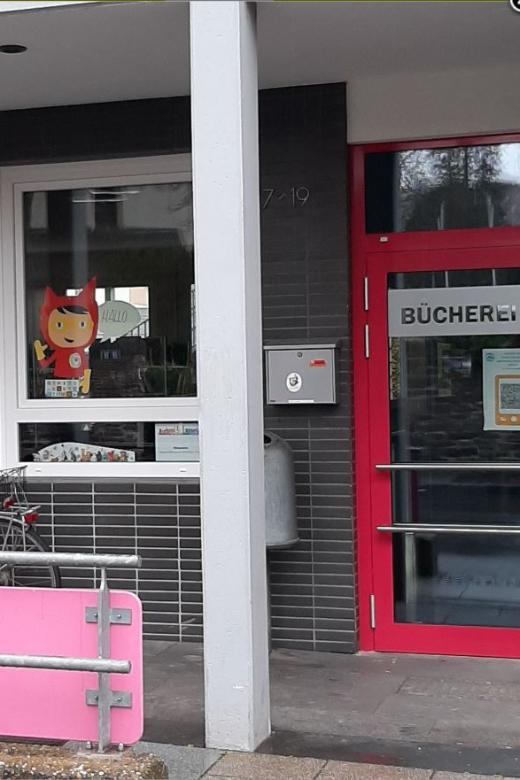 Blick auf die Eingangstür und 2 Fenster, davor steht ein Fahrrad. Ein Fenster ist mit dem Schriftzug der Bücherei beklebt