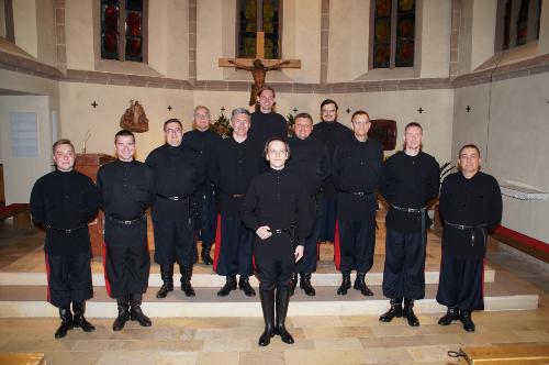 Zwölf Männer ganz in schwarz gekleidet stehen im Chorraum einer Kirche vor einem Kreuz
