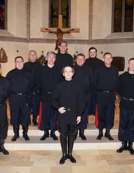 Zwölf Männer ganz in schwarz gekleidet stehen im Chorraum einer Kirche vor einem Kreuz