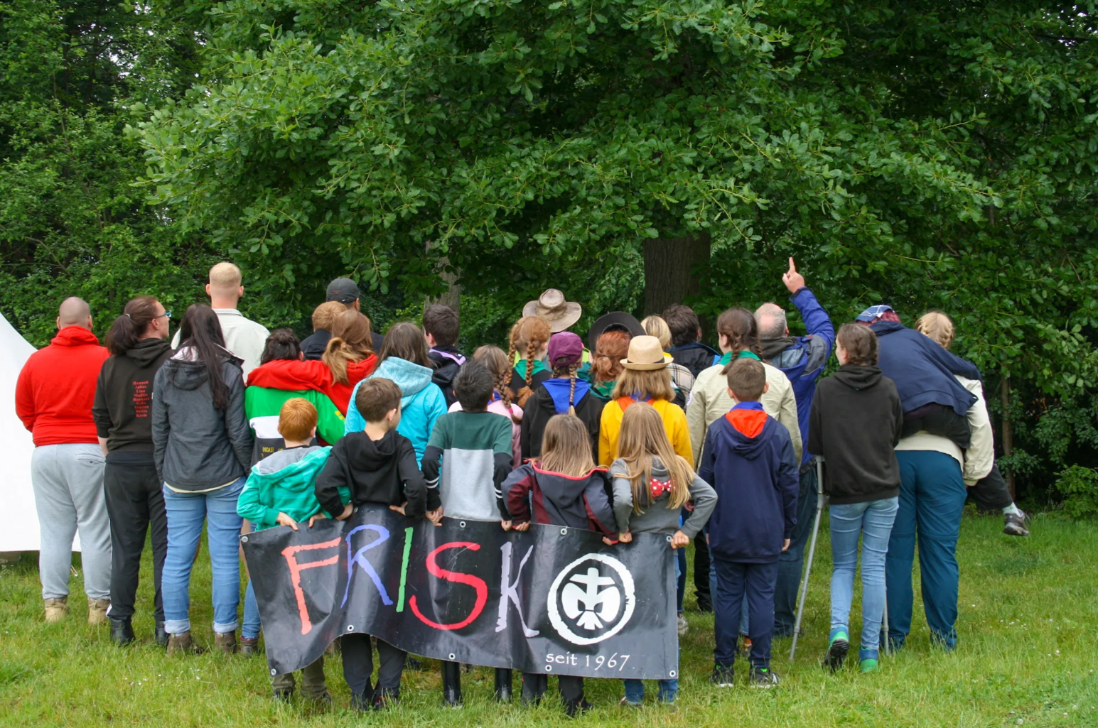 von hinten aufgenommenes Gruppenfoto des Pfadfinderstammes DSPG FRISKO, alle haben den Blick auf den Wald vor ihnen gerichtet und halten hinter sich das Banner FRISKO