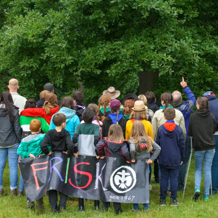 von hinten aufgenommenes Gruppenfoto des Pfadfinderstammes DSPG FRISKO, alle haben den Blick auf den Wald vor ihnen gerichtet und halten hinter sich das Banner FRISKO