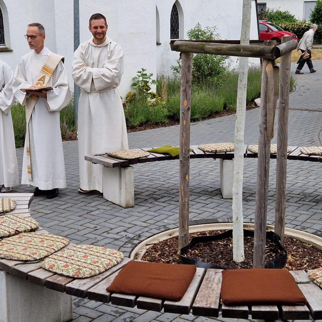Um einen kleinen Baum herum ist eine runde Bank mit Sitzkissen, im Hintergrund Diakon Wilbertz und zwei weitere Personen in lithurgischen Gewändern