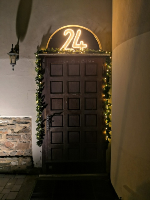 Fensterzauber (c) Julia Rosenbaum Über einer großen Holztür die Zahl 24, die leuchtet. Die Tür ist mit einer Tannengirlande und Lichterkette geschmückt