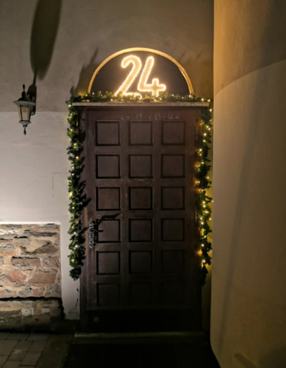 Über einer großen Holztür die Zahl 24, die leuchtet. Die Tür ist mit einer Tannengirlande und Lichterkette geschmückt