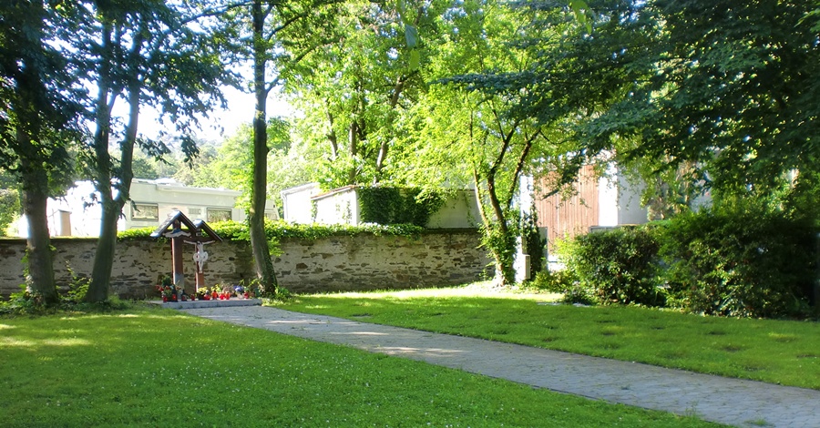 ein Weg auf dem Friedhof, am Ende eine Ablagefläche für Blumen mit zwei Kreuzen, rechts und links davon Rasenflächen mit eingelassenen Grabplatten. Im Hintergrund Bäume und eine Mauer