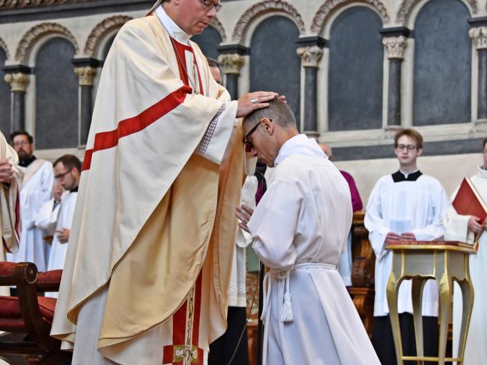 Heinz-Peter Wilbertz ist seitlich von hinten zu sehen. Er kniet auf Stufen vor dem Trierer Dom, bekleidet in eine weiße Kutte, vor ihm steht der Bischof , er hat seine Hände auf den Kopf gelegt