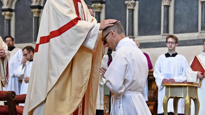 Heinz-Peter Wilbertz ist seitlich von hinten zu sehen. Er kniet auf Stufen vor dem Trierer Dom, bekleidet in eine weiße Kutte, vor ihm steht der Bischof , er hat seine Hände auf den Kopf gelegt