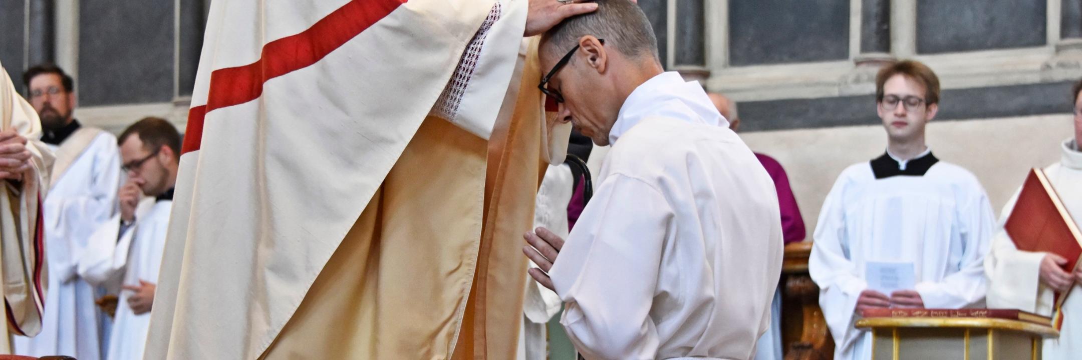 Heinz-Peter Wilbertz ist seitlich von hinten zu sehen. Er kniet auf Stufen vor dem Trierer Dom, bekleidet in eine weiße Kutte, vor ihm steht der Bischof , er hat seine Hände auf den Kopf gelegt