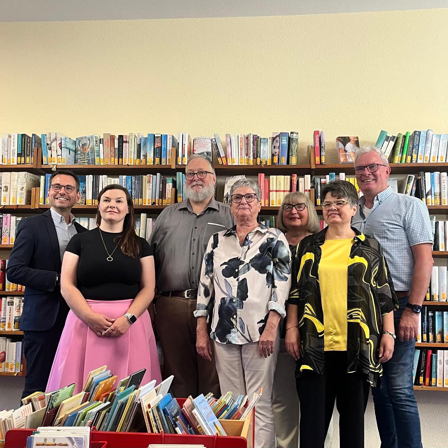 Feierlich gekleidete Personen vor Bücherregalen, davor Kisten mit Büchern