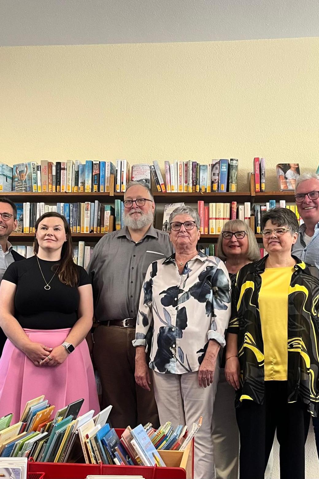 Feierlich gekleidete Personen vor Bücherregalen, davor Kisten mit Büchern