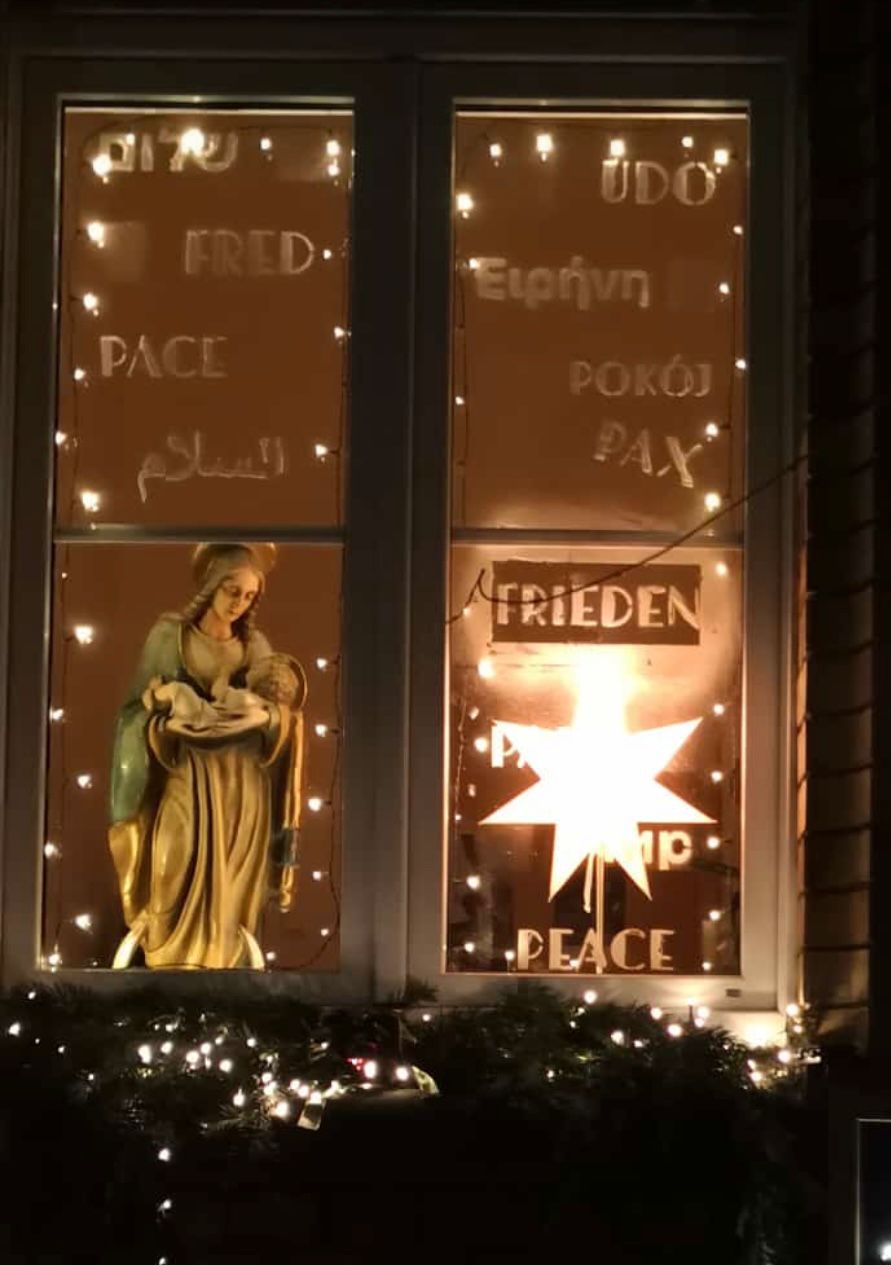 Zu sehen ist ein zweigeteiltes Fenster mit dem Wort Frieden in verschiedenen Sprachen umrankt von einer Lichterkette. Ferner ist im rechten Fensterflügel ein großer beleuchteter Stern, im linken Fensterflügel die Muttergottes mit dem Jesuskind zu sehen (c) Petra Gotthard / Pfarrei Koblenz St. Aposteln Zu sehen ist ein zweigeteiltes Fenster mit dem Wort Frieden in verschiedenen Sprachen umrankt von einer Lichterkette. Ferner ist im rechten Fensterflügel ein großer beleuchteter Stern, im linken Fensterflügel die Muttergottes mit dem Jesuskind zu sehen