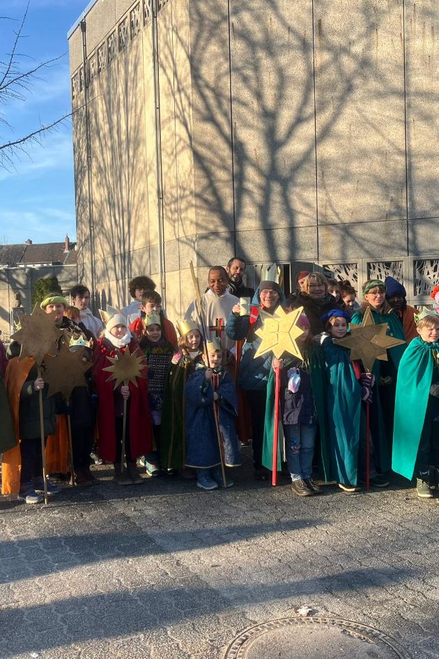 Eine Grupe Sternsinger mit ihren Betreuern und Pater Peter im Feierhof von St. Franziskus, im Hintergrund ist blauer Himmel und die Franziskuskirche zu sehen auf deren Betonwand Schatten  von  kahlen Bäumen zu sehen sind.