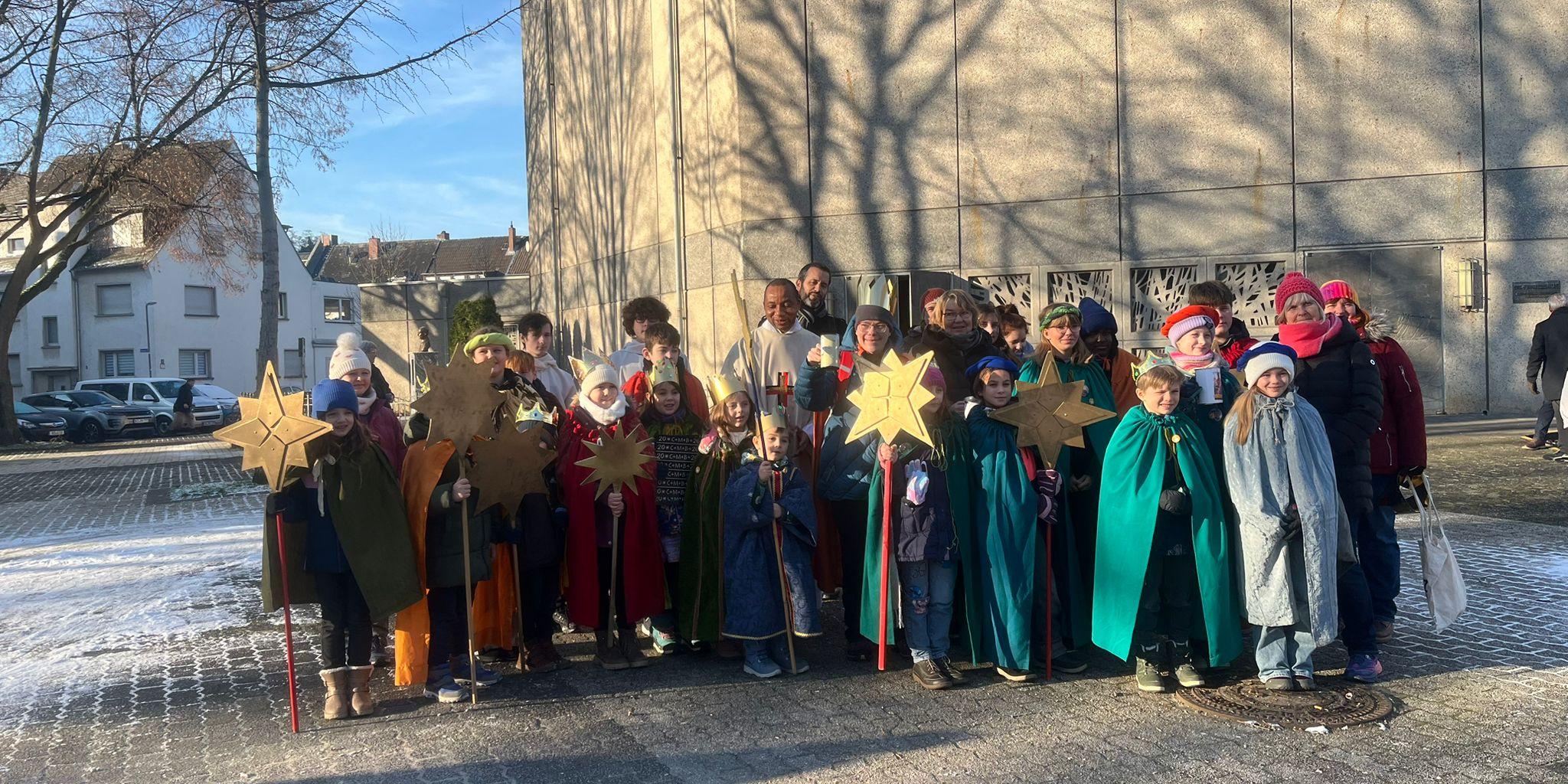 Eine Grupe Sternsinger mit ihren Betreuern und Pater Peter im Feierhof von St. Franziskus, im Hintergrund ist blauer Himmel und die Franziskuskirche zu sehen auf deren Betonwand Schatten  von  kahlen Bäumen zu sehen sind.