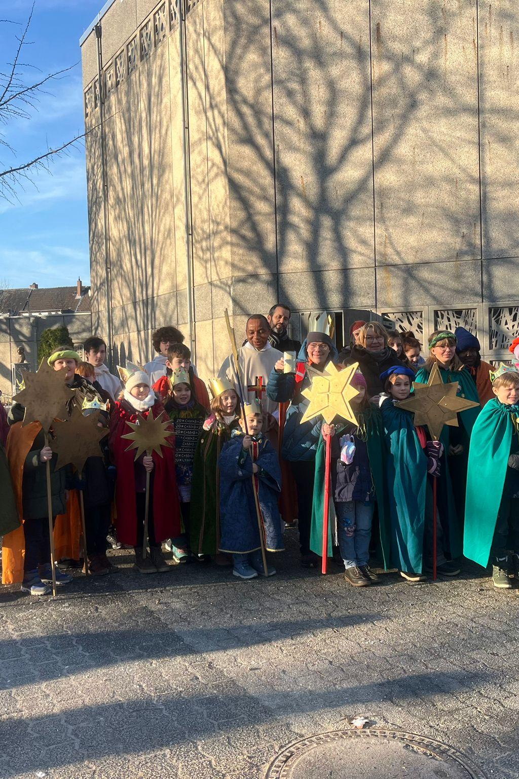Eine Grupe Sternsinger mit ihren Betreuern und Pater Peter im Feierhof von St. Franziskus, im Hintergrund ist blauer Himmel und die Franziskuskirche zu sehen auf deren Betonwand Schatten  von  kahlen Bäumen zu sehen sind.