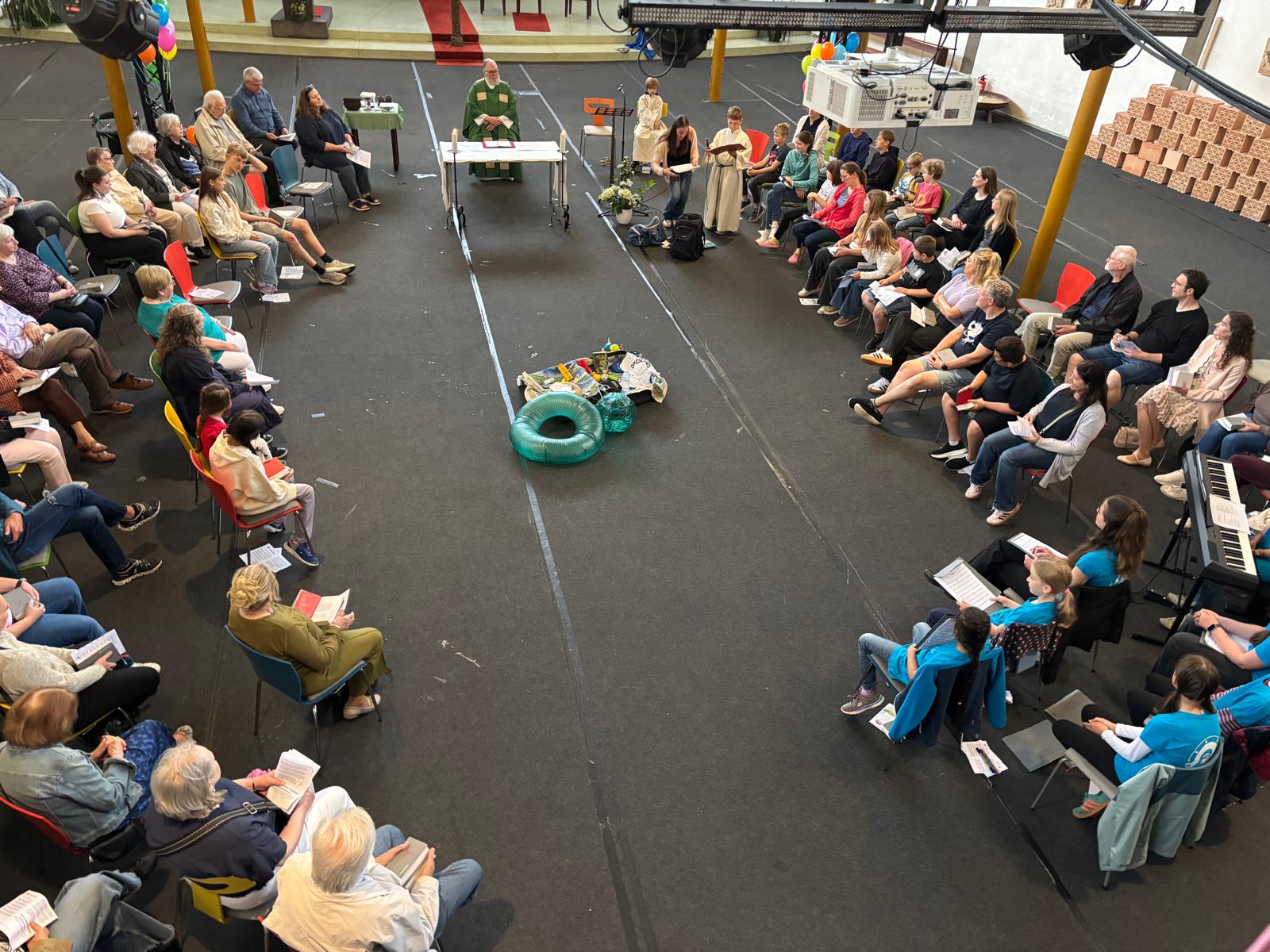 Gottesdienstbesucher sitzen rechts und links von einem Altartisch im Kreis. In der Mitte ein offener Koffer, Schwimmreif, Ball (c) Petra Gotthard / Pfarrei Koblenz St. Aposteln Gottesdienstbesucher sitzen rechts und links von einem Altartisch im Kreis. In der Mitte ein offener Koffer, Schwimmreif, Ball