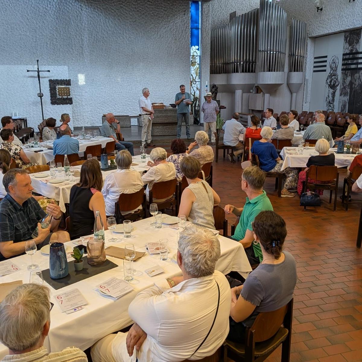 An sechs großen mit Wein-, Wassergläsern und Tellern eingedeckten Tischen sitzen Teilnehmer der Veranstaltung Wein und Brot und lauschen den Referenten