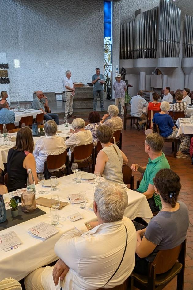 An sechs großen mit Wein-, Wassergläsern und Tellern eingedeckten Tischen sitzen Teilnehmer der Veranstaltung Wein und Brot und lauschen den Referenten