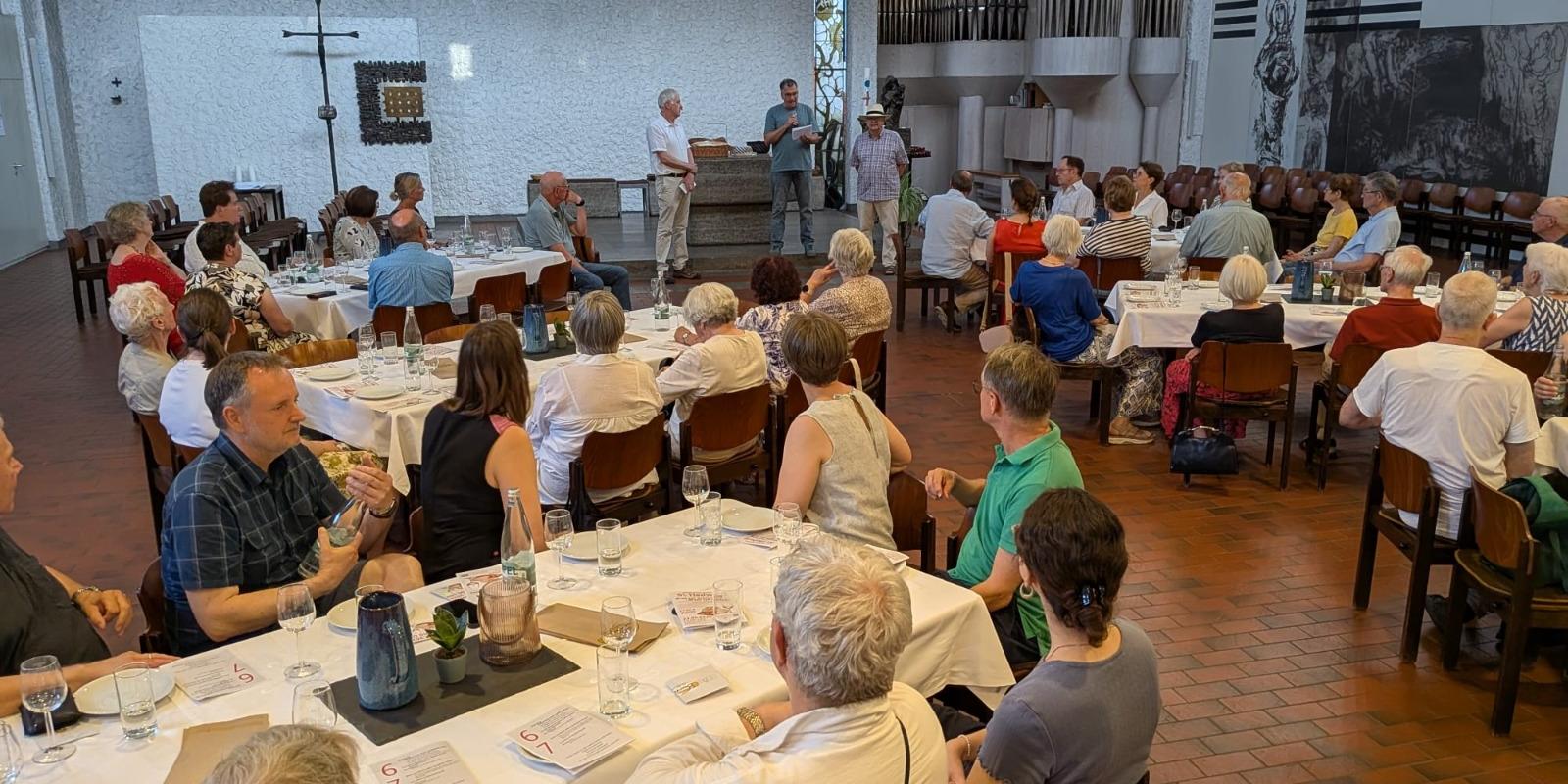 An sechs großen mit Wein-, Wassergläsern und Tellern eingedeckten Tischen sitzen Teilnehmer der Veranstaltung Wein und Brot und lauschen den Referenten