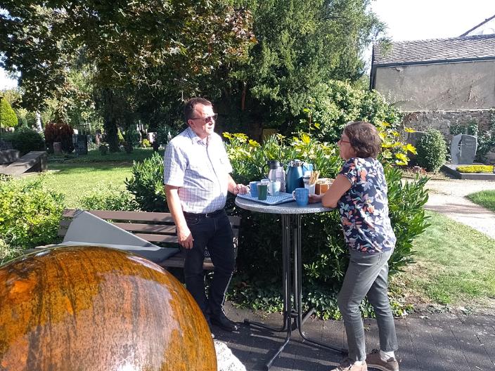 Ein Mann und eine Frau stehen an einem Stehtisch im Hintergrund ein Friedhof im Vordergrund ein Brunnen
