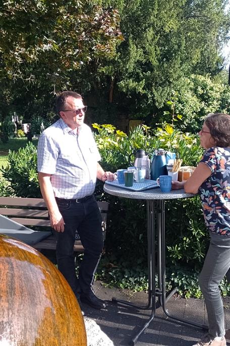 Ein Mann und eine Frau stehen an einem Stehtisch im Hintergrund ein Friedhof im Vordergrund ein Brunnen