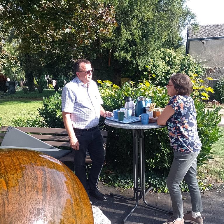 Ein Mann und eine Frau stehen an einem Stehtisch im Hintergrund ein Friedhof im Vordergrund ein Brunnen