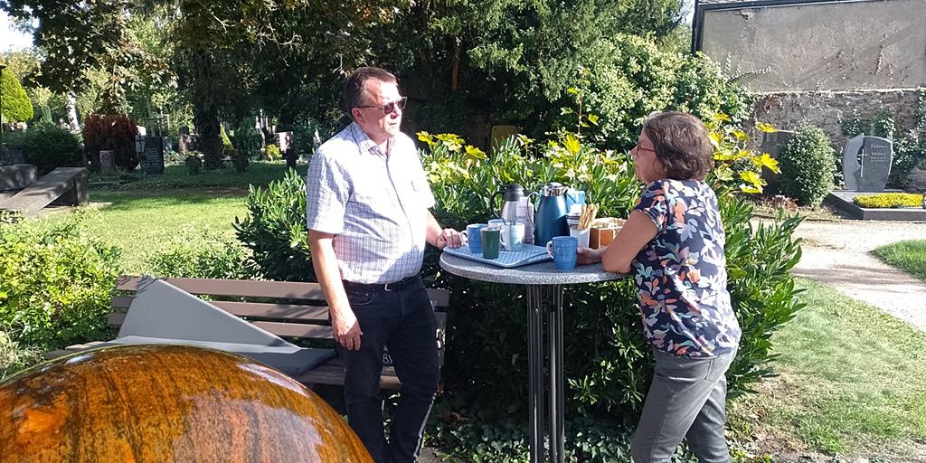 Ein Mann und eine Frau stehen an einem Stehtisch im Hintergrund ein Friedhof im Vordergrund ein Brunnen