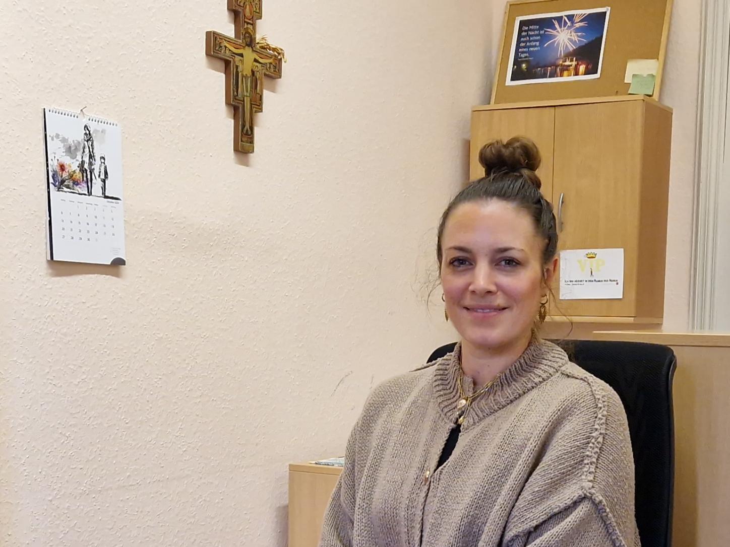 zu sehen ist Janine Theisen, sitzend auf einem Bürostuhl, im Hintergrund eine Wand mit einem Kreuz