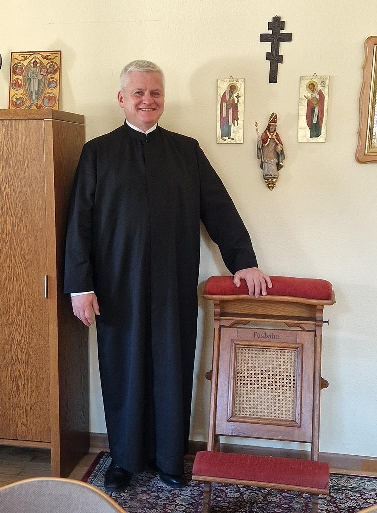 Jörg Stein ist in Ganzkörperaufnahme zu sehen, im liturgischen Gewand, eine Hand liegt auf einem Beichtstuhl, er lächelt