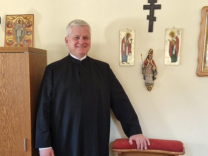 Jörg Stein ist in Ganzkörperaufnahme zu sehen, im liturgischen Gewand, eine Hand liegt auf einem Beichtstuhl, er lächelt