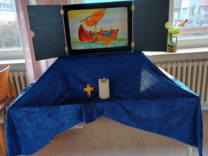 Auf einem Tisch ist ein mit einem blauen Tuch überdeckter Sockel auf dem ein Kamishibaitheater aufgebaut ist. Davor steht ein Kreuz und eine Kerze